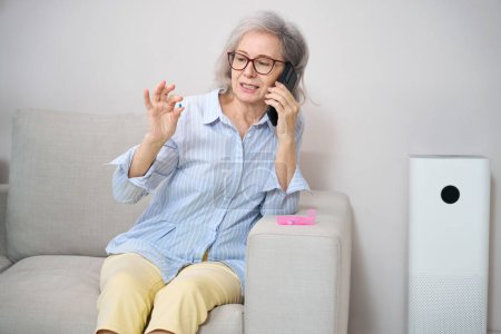 Foto de Pensionista sostiene una píldora en sus manos y habla por teléfono, ella se sienta en un cómodo sofá - Imagen libre de derechos