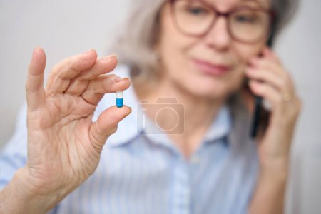 Foto de Mujer mayor sostiene una píldora en sus manos y habla por teléfono, tiene el pelo gris - Imagen libre de derechos