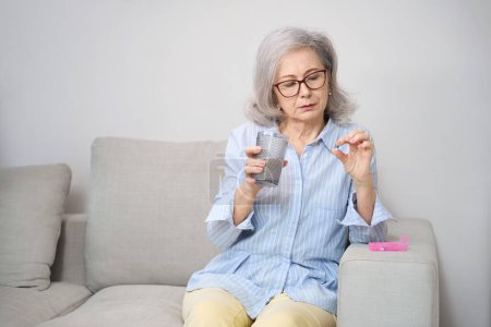 Foto de Bonito pensionista sentado en un sofá con un vaso de agua para beber una píldora, un pillbox se encuentra cerca - Imagen libre de derechos