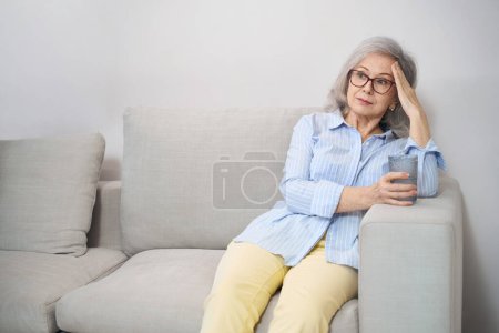 Foto de Mujer bastante jubilada sentada en el sofá con un vaso de agua, tiene un poco de dolor de cabeza - Imagen libre de derechos