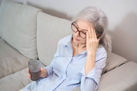 Téléchargez les photos : Grand-mère aux cheveux gris souffre d'un mal de tête, elle s'assoit sur le canapé avec un verre d'eau - en image libre de droit