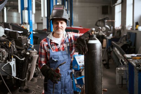 Téléchargez les photos : Employé d'un atelier de réparation automobile se tient dans un atelier avec machine à souder, un homme appuyé sur une bouteille de gaz - en image libre de droit