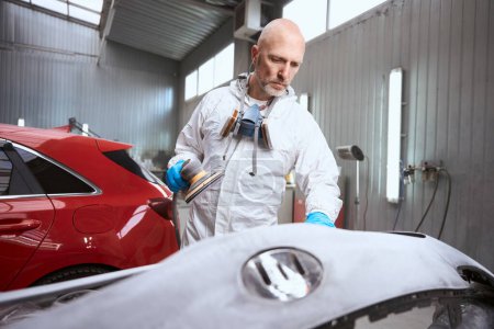 Photo for Auto repair shop employee grinds with unpainted car bumper, man in protective overalls - Royalty Free Image