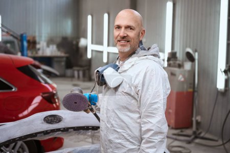 Photo pour Homme avec un respirateur autour de son cou tient la machine pour meuler la surface de l'automobile dans ses mains, en arrière-plan voiture rouge - image libre de droit