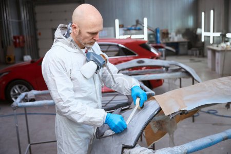 Photo pour L'ouvrier d'atelier de réparation automobile prépare le pare-chocs de voiture pour la peinture, l'homme utilise du ruban adhésif - image libre de droit