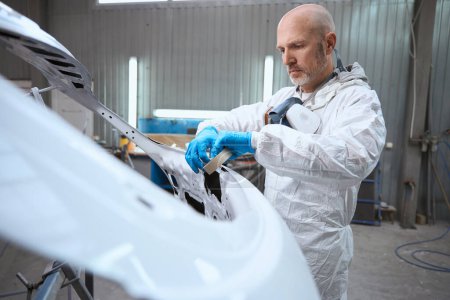 Photo for Employee of the paint shop prepares part of car body for work, the man has a respirator and protective gloves - Royalty Free Image