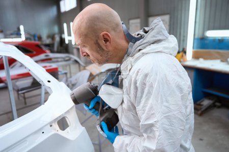 Foto de Trabajador de taller de reparación de automóviles que utiliza aerosol pulverizador para pintar la carrocería del coche, hombre en mono protector - Imagen libre de derechos