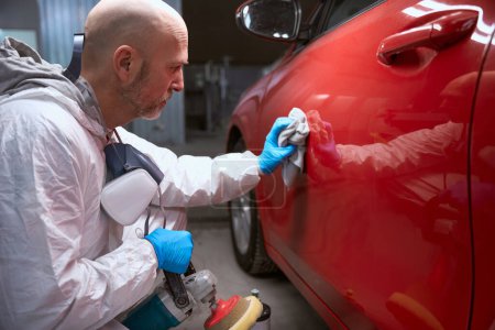 Photo pour Contremaître d'âge moyen essuie le corps d'une voiture rouge avec un chiffon doux, un homme en salopette de travail - image libre de droit