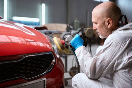 Téléchargez les photos : L'homme dans un atelier de réparation broie le phare d'une voiture rouge, un homme utilise un outil spécial - en image libre de droit