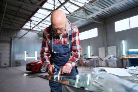 Téléchargez les photos : Travailleur masculin dans un atelier de réparation travaille avec un pare-brise, il utilise un outil spécial - en image libre de droit