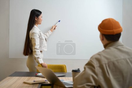 Téléchargez les photos : Joyeuse employée de bureau pointant avec un marqueur au tableau blanc vers son collègue assis au bureau - en image libre de droit