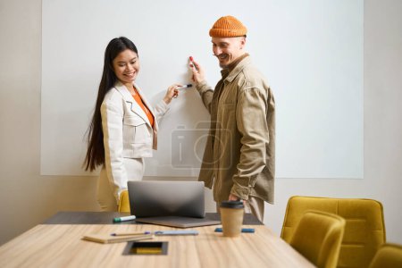 Foto de Smiling empleados de la compañía señalando con marcadores en la pizarra blanca mientras mira la pantalla del ordenador portátil - Imagen libre de derechos
