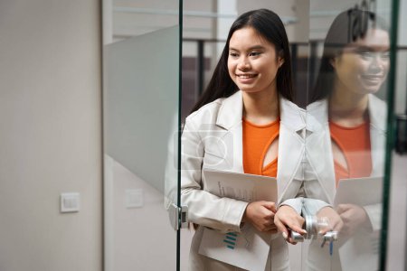 Téléchargez les photos : Portrait taille haute d'une femme d'affaires asiatique heureuse avec pile de documents dans les mains ouvrant une porte en verre - en image libre de droit