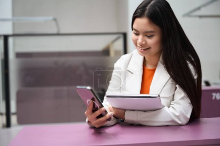 Photo for Joyful Asian corporate worker with portable computer in hand looking at cellphone touch screen - Royalty Free Image