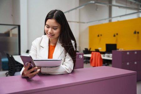 Téléchargez les photos : Jeune employée souriante avec ordinateur portable à la main regardant l'écran tactile du smartphone - en image libre de droit