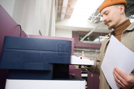 Téléchargez les photos : Travailleur d'entreprise concentré sérieux avec des feuilles de papier à la main touchant panneau de commande de photocopieur - en image libre de droit
