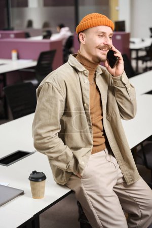 Foto de Sonriente trabajador corporativo feliz sentado en el escritorio mientras habla por teléfono celular en una oficina abierta - Imagen libre de derechos
