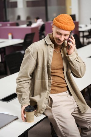 Foto de Agradable empleado de la compañía que busca taza de papel durante la conversación telefónica mientras está sentado en el escritorio - Imagen libre de derechos