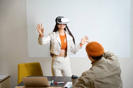 Photo for Pleased corporate worker in VR glasses standing at office desk in front of her coworker - Royalty Free Image
