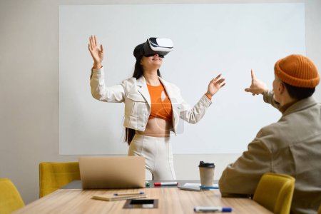 Photo for Smiling employee in virtual reality glasses standing at desk before her male colleague - Royalty Free Image