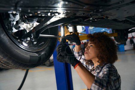 Foto de Joven mecánico de automóviles realiza el mantenimiento del servicio de coche en un taller de reparación de automóviles, la mujer inspecciona el coche desde abajo - Imagen libre de derechos