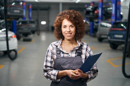 Photo pour Femme souriante mécanicien automobile se tient au milieu d'un atelier de réparation automobile, elle a dossier bleu dans ses mains - image libre de droit