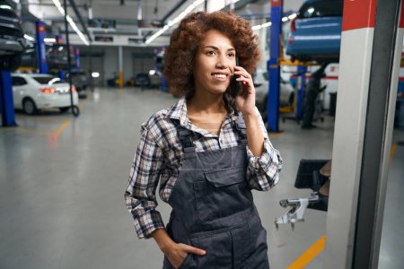 Photo for Woman auto mechanic in a car repair shop talking on a mobile phone, a repairwoman in a plaid shirt - Royalty Free Image