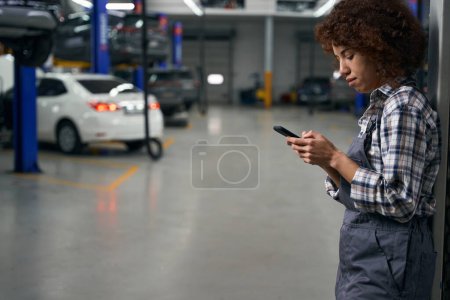 Photo pour Employé multiracial d'un atelier de réparation automobile moderne appuie sur les boutons de téléphone mobile, réparatrice dans une chemise à carreaux - image libre de droit