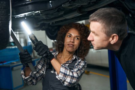 Photo pour Mécanicien féminin multiracial et client inspectant la voiture d'en bas, la femme a une lampe spéciale - image libre de droit