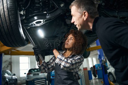 Foto de Mujer rizada mecánico de automóviles y el cliente inspeccionar el coche desde abajo, la mujer tiene lámpara especial - Imagen libre de derechos