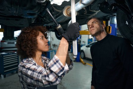 Photo pour Jeune femme mécanicien automobile et un client inspecter la voiture d'en bas, la femme a une lampe spéciale - image libre de droit