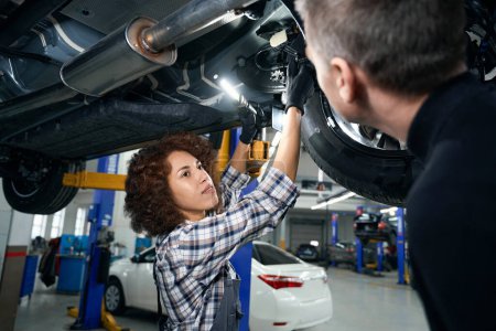 Photo pour Femme mécanicien automobile en salopette de travail et un client inspecter une voiture d'en bas, autour des voitures modernes - image libre de droit