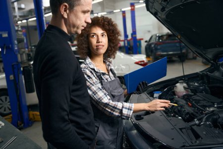 Foto de Hombre con ropa casual se comunica con un capataz en un taller de reparación de automóviles, una mujer multirracial tiene una carpeta azul - Imagen libre de derechos