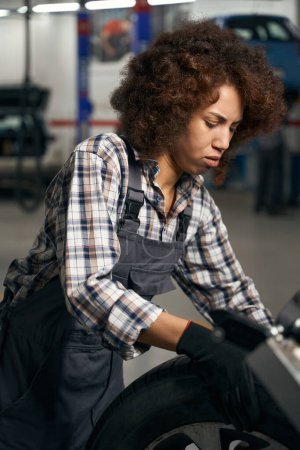 Foto de Mujer joven especialista en un taller de reparación de automóviles trabaja con una rueda de coche, mujer en mono de trabajo - Imagen libre de derechos