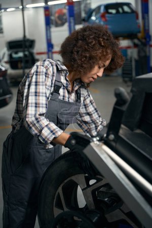 Photo pour Femme dans un atelier de réparation automobile travaille avec une roue de voiture, femme en salopette de travail - image libre de droit