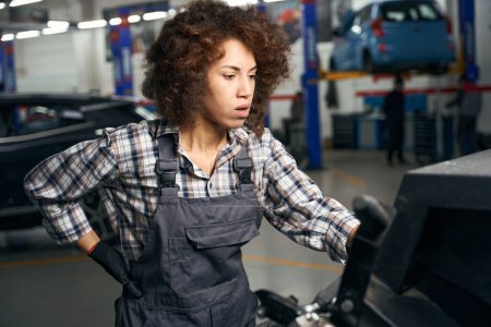 Photo for Pretty multiracial woman is working in atomist shop at her workplace, in background the car is fixed on a lift - Royalty Free Image