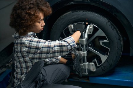 Téléchargez les photos : Jeune femme aux cheveux bouclés ajuste l'alignement des roues dans l'atelier de réparation automobile, contremaître en vêtements de travail - en image libre de droit