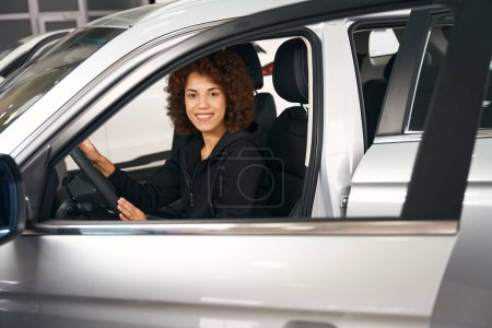 Photo for Smiling multiracial female shopper sits behind the wheel in the salon of brand new car, she chooses the right model - Royalty Free Image