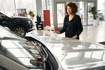 Foto de Sonriente mujer multirracial se encuentra cerca de un coche nuevo, el coche se vende en un salón especializado - Imagen libre de derechos