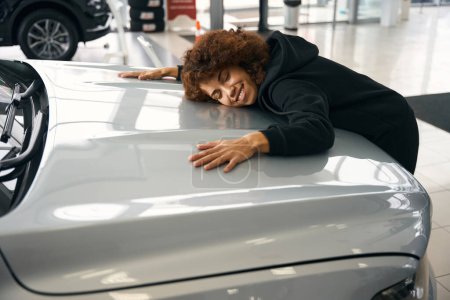 Téléchargez les photos : Heureuse femme multiraciale serrant le capot d'une nouvelle voiture, la voiture est vendue dans un salon spécialisé - en image libre de droit