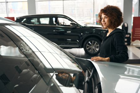 Photo for Smiling multiracial woman put her hand on the hood of a new car, the car is sold in specialized salon - Royalty Free Image