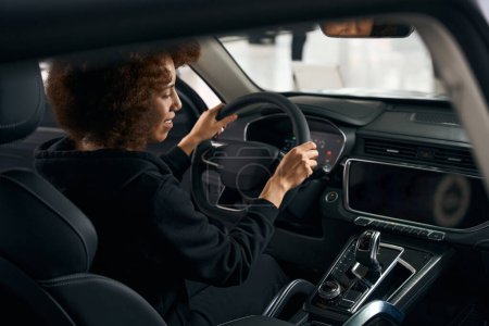 Photo for Smiling multiracial female shopper sits behind the wheel in the interior of a modern car, woman in comfortable clothes - Royalty Free Image