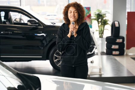 Photo for Happy multiracial car dealership shopper holding new car keys in her hands, woman in velvet casual wear - Royalty Free Image