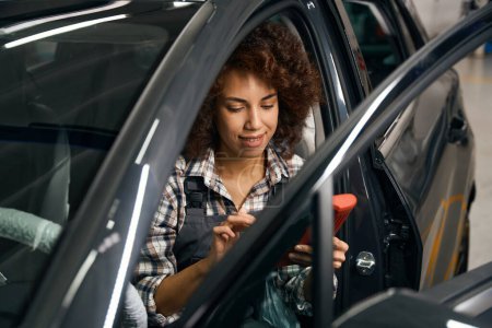 Téléchargez les photos : Femme multiraciale mécanicien automobile est assis dans une voiture avec un dispositif de diagnostic informatique, volant à l'abri de la pollution - en image libre de droit
