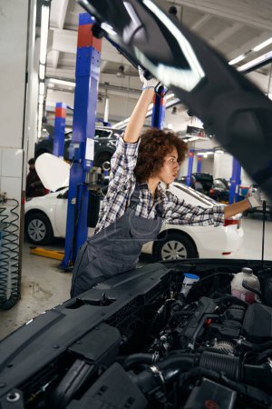 Foto de Bastante taller de reparación de automóviles empleado levanta la capucha del coche, mujer con guantes de trabajo - Imagen libre de derechos