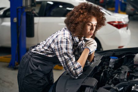Foto de Mujer reflexiva mecánico de automóviles inspecciona el coche bajo el capó, el coche está siendo atendido - Imagen libre de derechos
