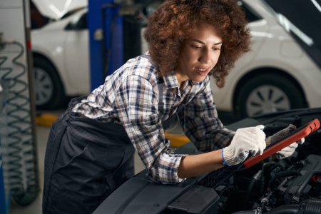 Téléchargez les photos : Femme technicienne en vêtements de travail effectue le diagnostic informatique d'une voiture, une femme en gants de protection - en image libre de droit