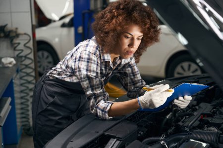Téléchargez les photos : Femme technicienne en vêtements de travail se tient près d'une voiture avec une capuche ouverte, elle fixe les pannes dans un dossier bleu - en image libre de droit