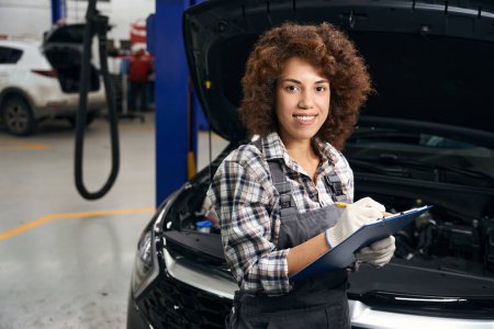 Photo for Young female technician stands near the machine in the workshop, she writes something in a blue folder - Royalty Free Image
