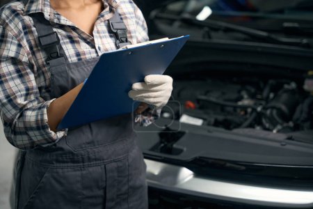 Foto de Técnica en ropa de trabajo está cerca de un coche en un taller, escribe algo en una carpeta azul - Imagen libre de derechos